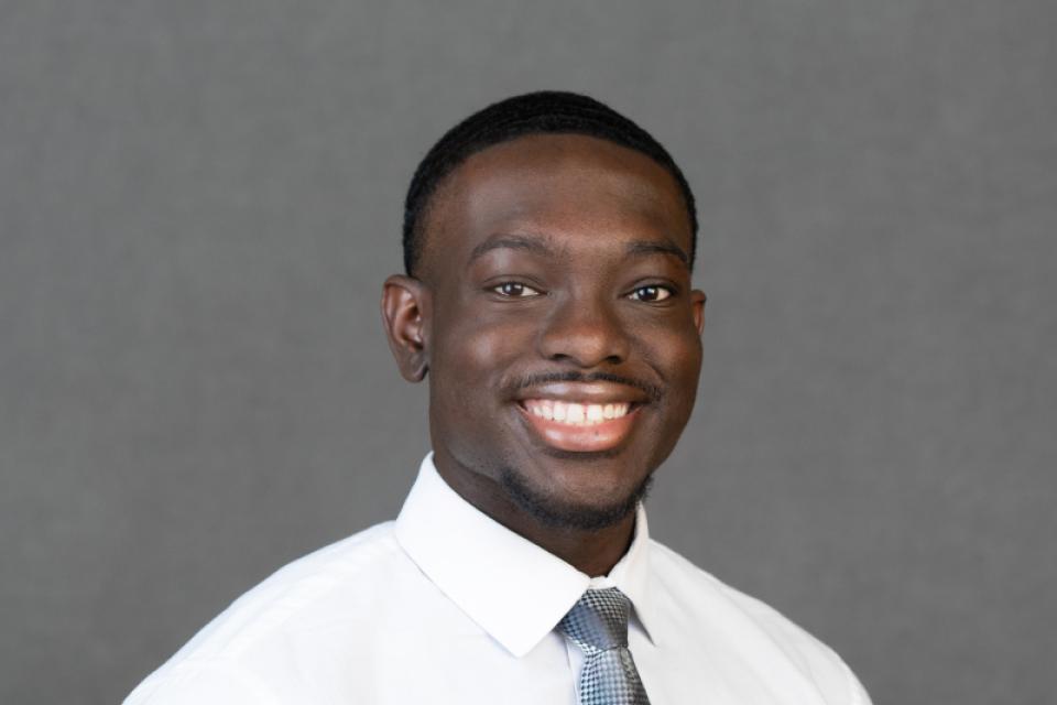 A man wearing a white dress shirt and patterned tie smiles at the camera against a plain gray background.