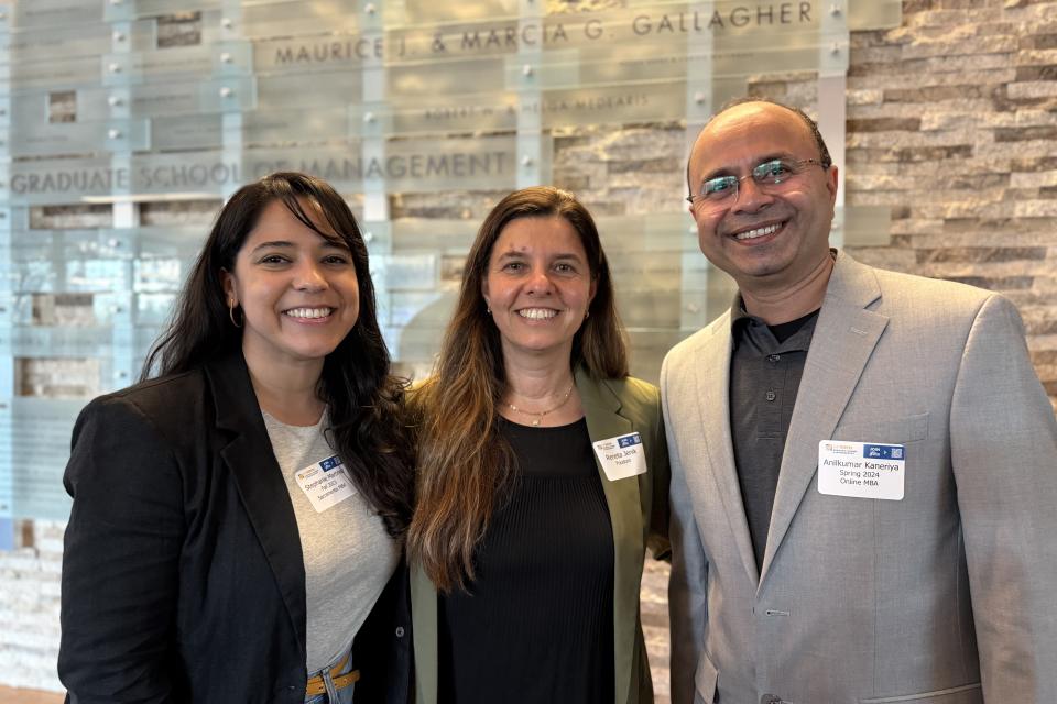 Three people stand indoors, smiling at the camera. They wear business attire and conference name tags. A wall sign behind them reads "Graduate School of Management.