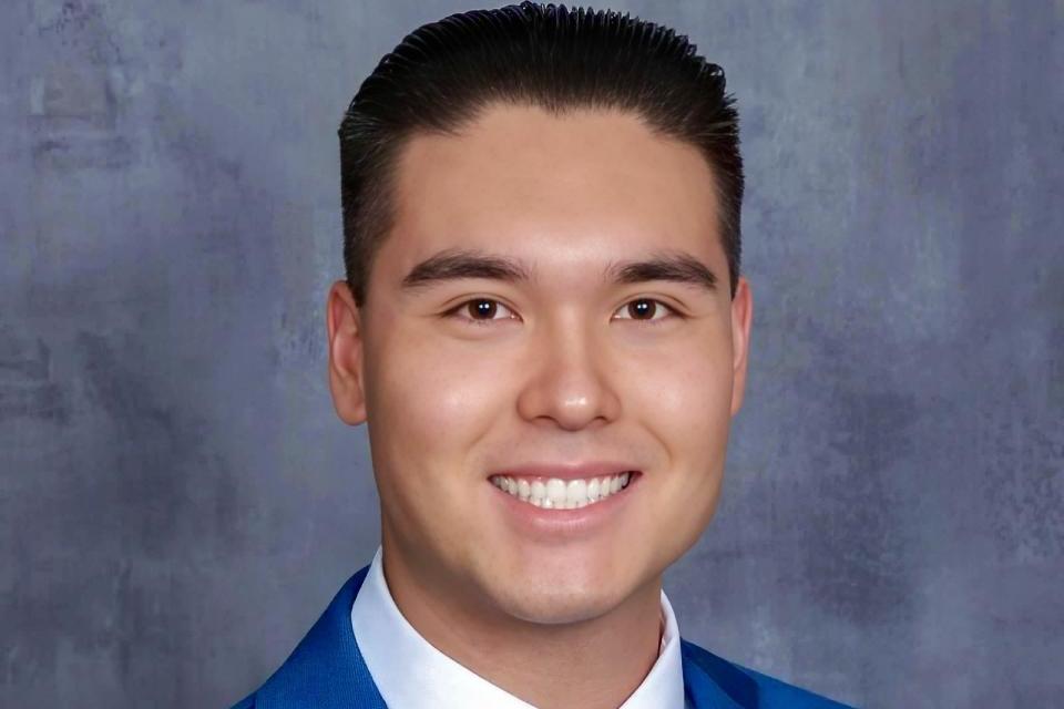 A young man wearing a blue suit, white shirt, and striped tie smiles in front of a gray mottled background.