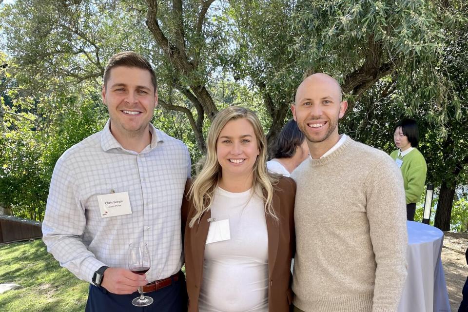 Three people stand outdoors, smiling at the camera. The two men wear business casual attire; the woman wears a white top and brown blazer. Trees and other event attendees are in the background.