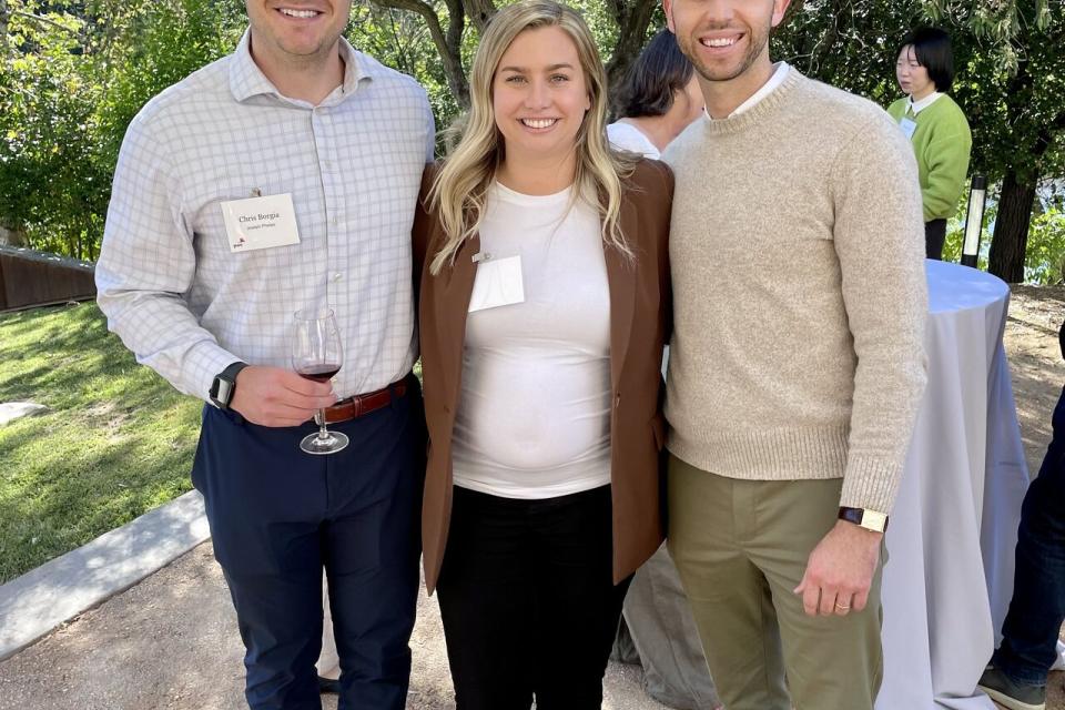 Three people stand outdoors, smiling at the camera. The two men wear business casual attire; the woman wears a white top and brown blazer. Trees and other event attendees are in the background.