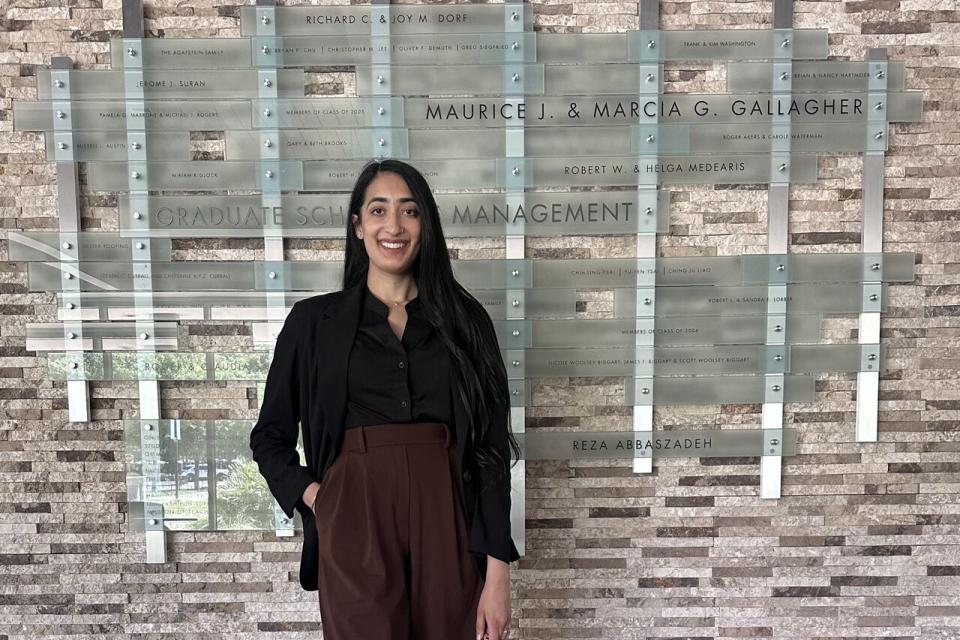 A woman stands indoors in business attire, holding a handbag and smiling, in front of a wall displaying names and a sign reading “Graduate School of Management.”