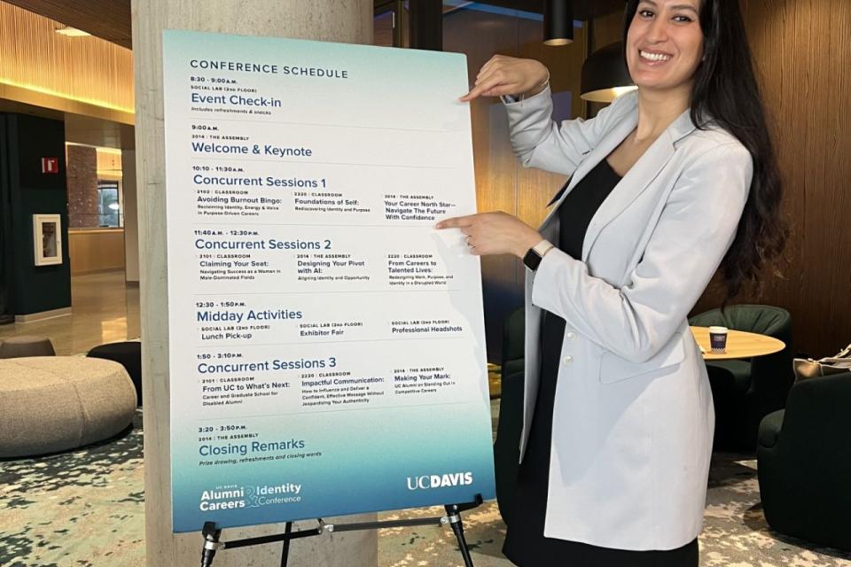 A woman in a light blazer stands indoors, smiling and pointing at a large printed conference schedule on an easel at a UC Davis event.