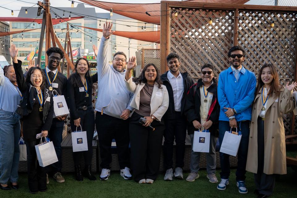 A group of eleven people pose together outdoors, some holding gift bags and wearing medals, smiling and raising their hands in celebration.