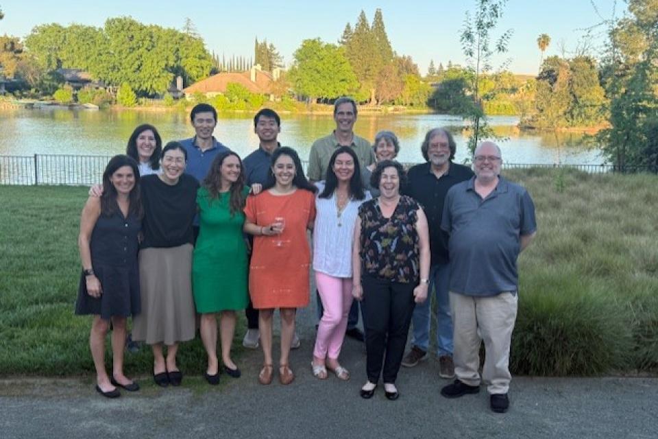 13 professors at the conference standing outdoors in front of a lake, smiling at the camera on a sunny day.