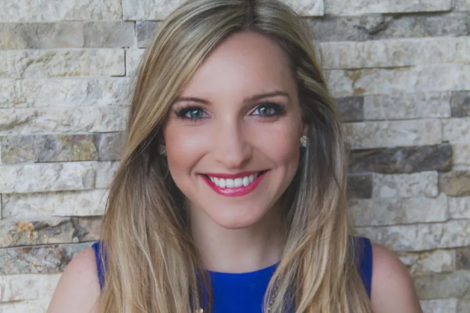 A woman with long blonde hair, wearing a blue sleeveless top and a floral necklace, smiles in front of a textured stone wall background.