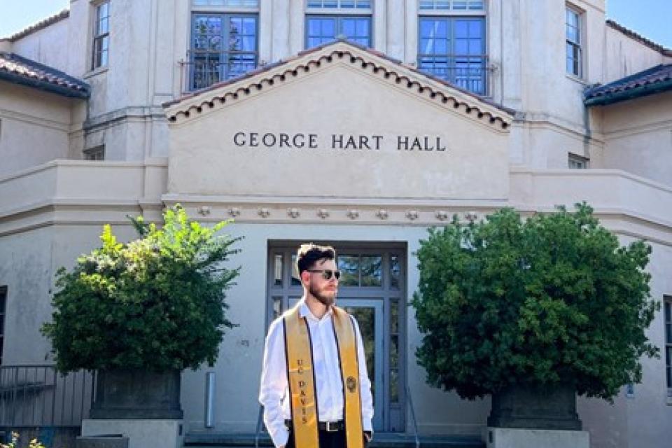 A man wearing a graduation stole stands in front of George Hart Hall on a sunny day.