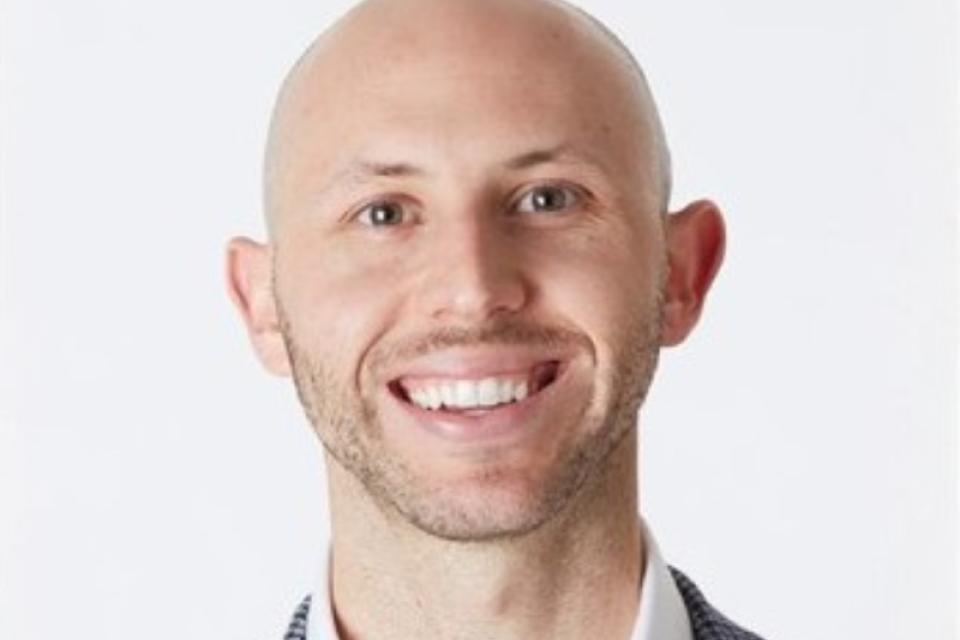 A smiling bald man wearing a white shirt and a checkered blazer, posed against a plain white background.