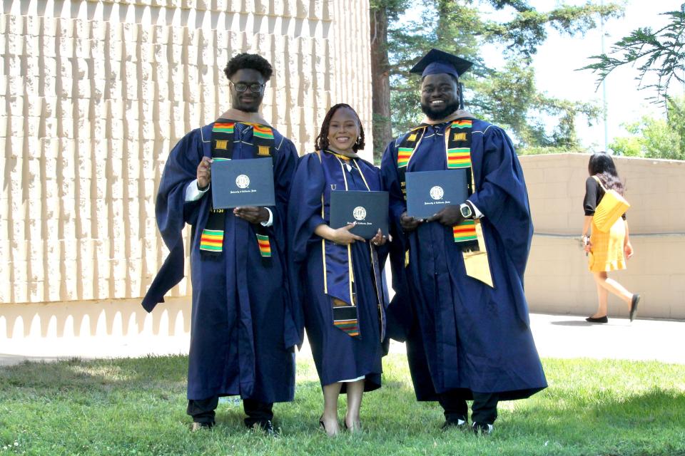Three graduates in navy blue caps and gowns hold diplomas and smile outdoors; another person walks in the background.