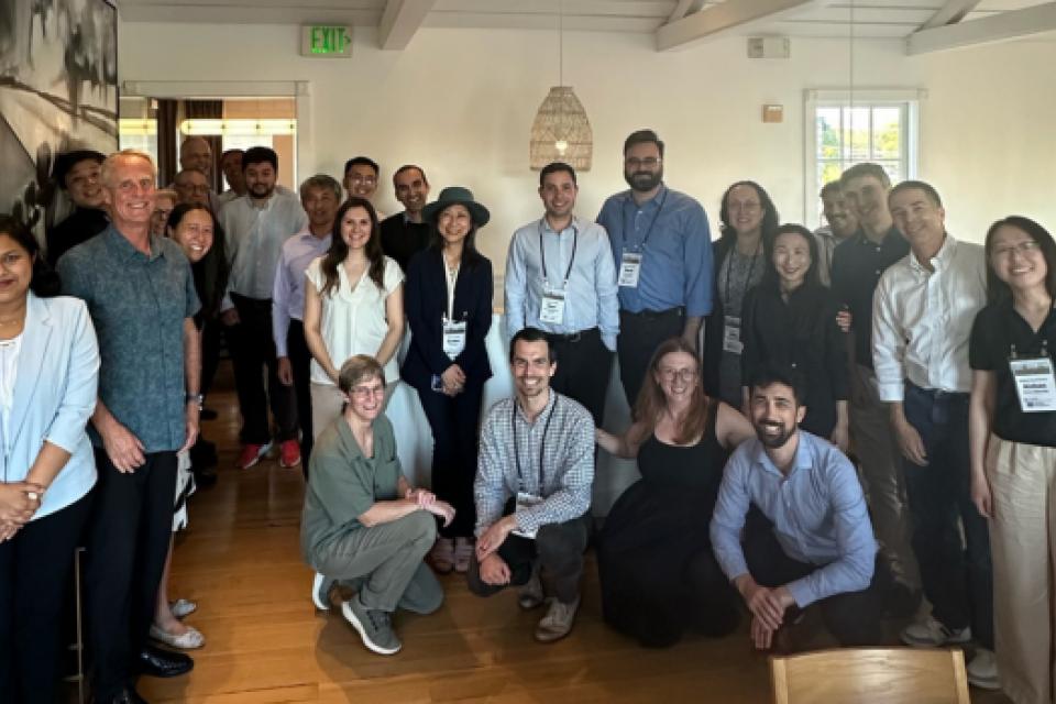 A group of about 20 people pose together indoors, some standing and some kneeling, in a well-lit room with wooden floors and white walls.