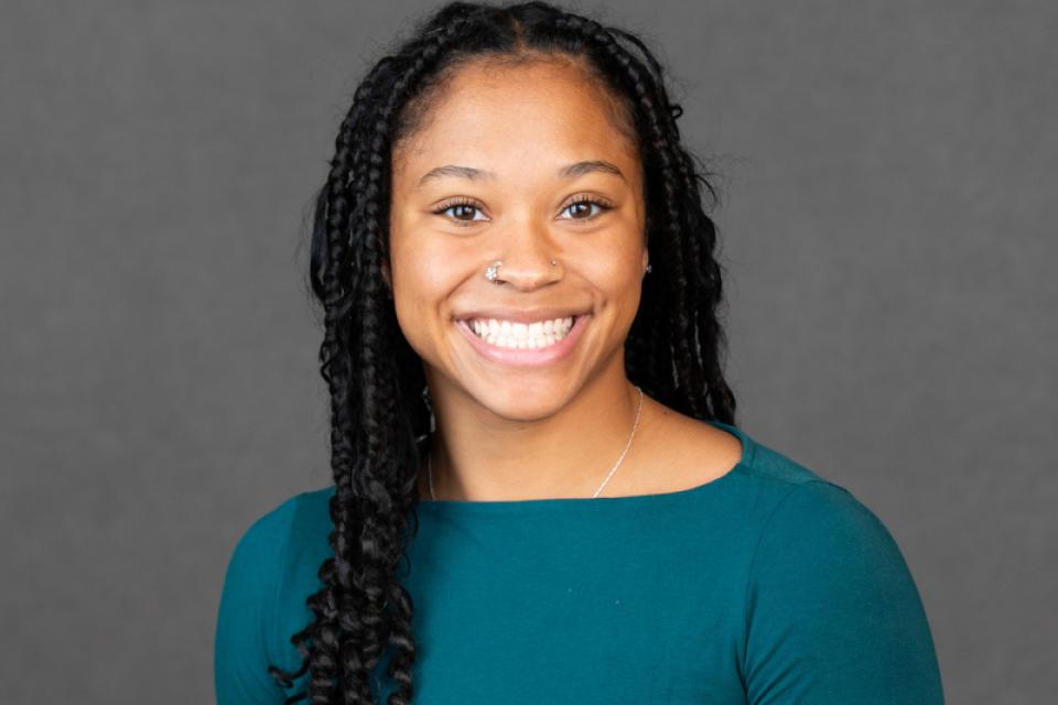 A woman with long braided hair, wearing a teal top, smiles at the camera against a plain gray background.
