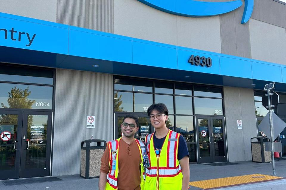 Two people wearing safety vests stand in front of an Amazon building entrance with the Amazon logo and address 4930 visible above them.