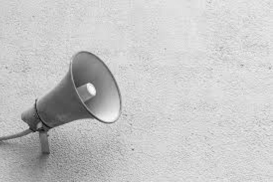 A wall-mounted megaphone or loudspeaker is shown against a plain, textured wall in black and white.