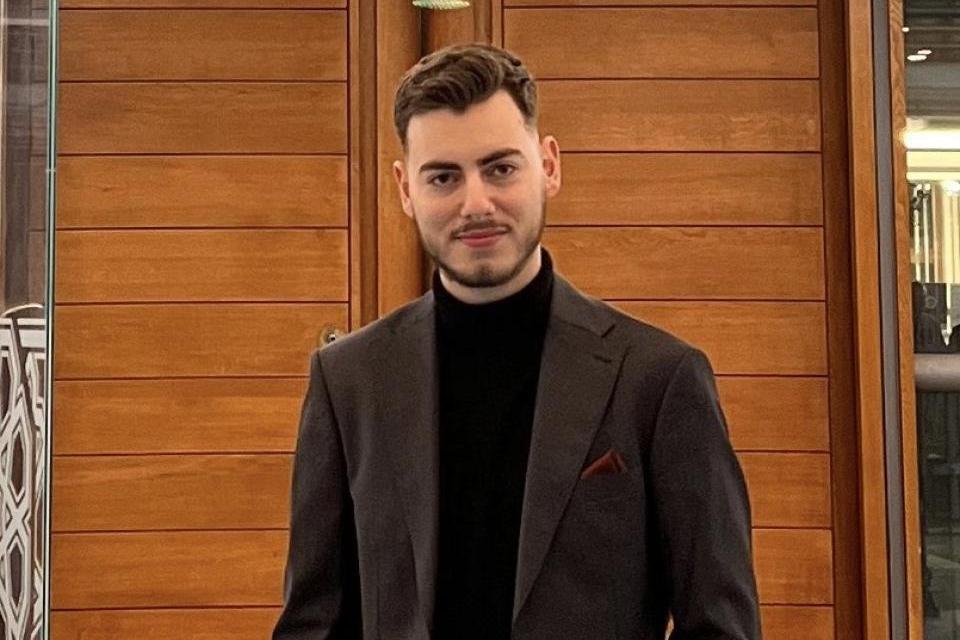 A man in a dark suit and black turtleneck stands indoors in front of wooden panel doors, with his hands in his pockets, facing the camera.