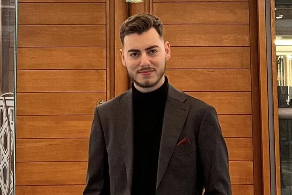 A man in a dark suit and black turtleneck stands indoors in front of wooden panel doors, with his hands in his pockets, facing the camera.