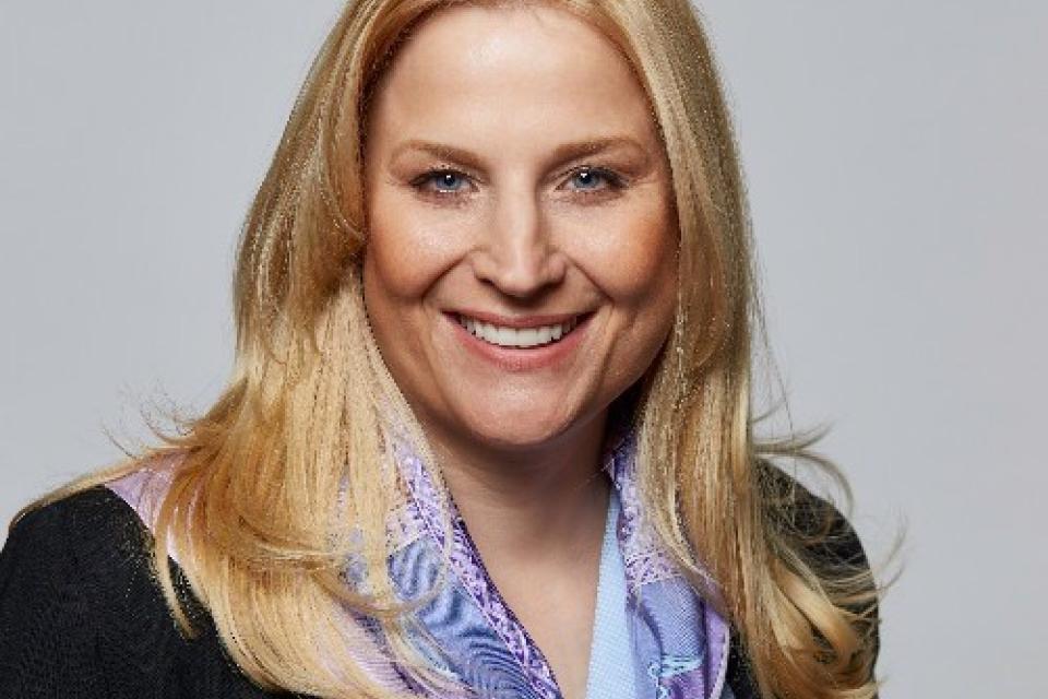 A woman with long blonde hair smiles, wearing a black blazer and a light purple patterned scarf, against a plain gray background.