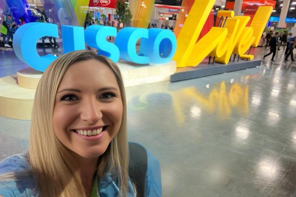 A woman takes a selfie in front of a large "CISCO Live" sign at a convention or expo, with colorful decorations and other attendees in the background.