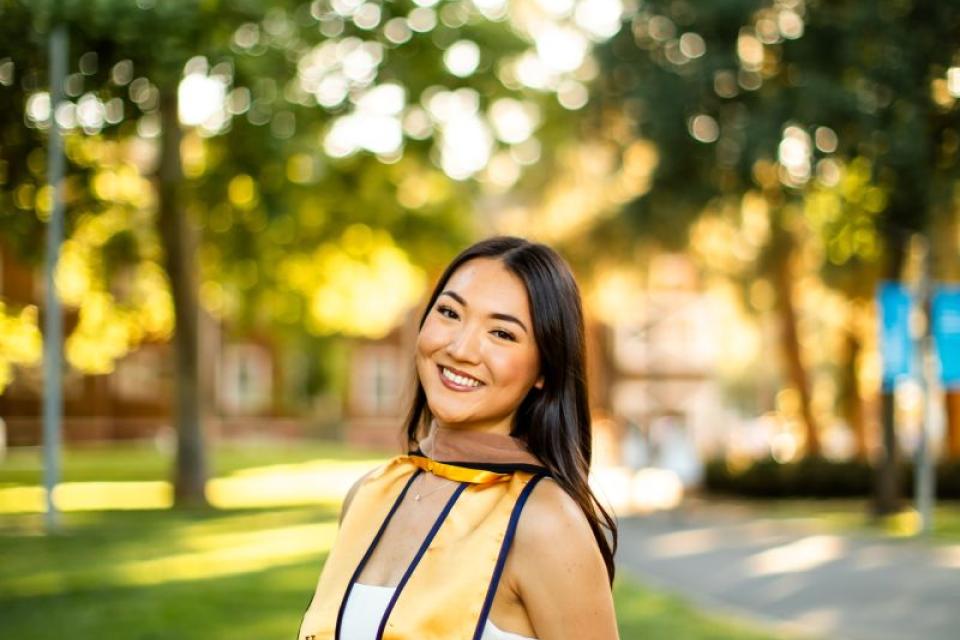 A young woman in a graduation stole and white dress stands outdoors by a brick wall, smiling, with trees and sunlight in the background.