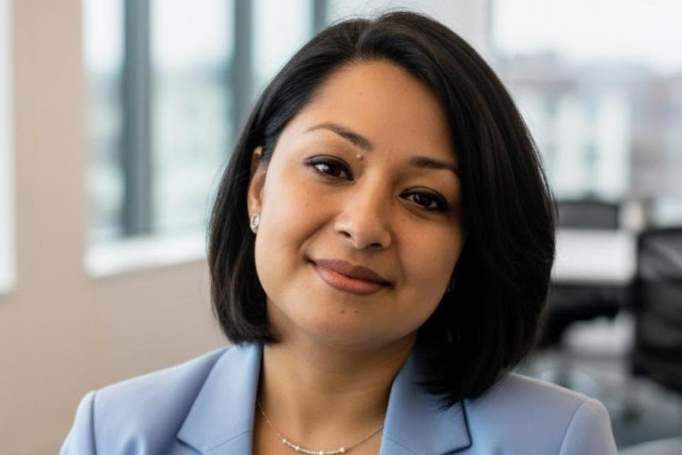 A woman with short dark hair wearing a light blue blazer and white top sits in a modern office with large windows.