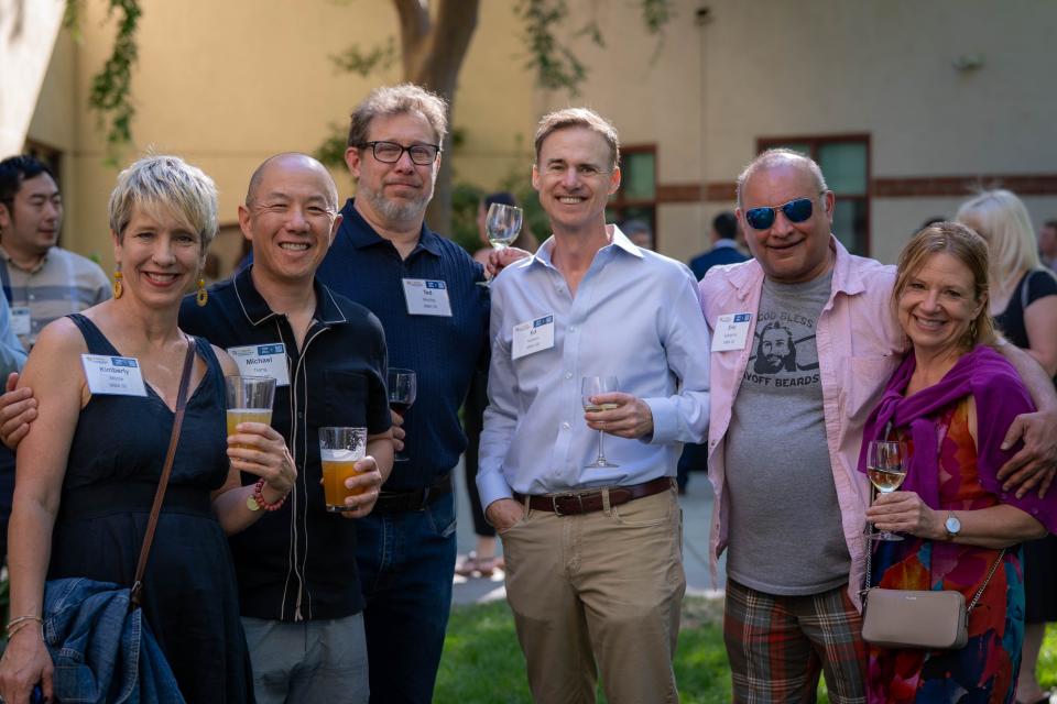 A group of six adults, wearing name tags and holding drinks, pose together and smile at an outdoor social gathering.