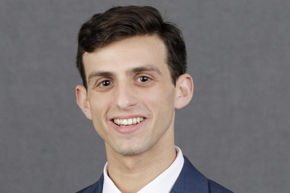 Jacob Renneisen wearing a blue suit, white shirt, and red tie is smiling in front of a plain gray background.