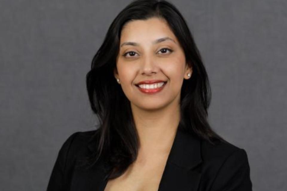 A woman with long dark hair wearing a black blazer and blue top smiles in front of a plain gray background.