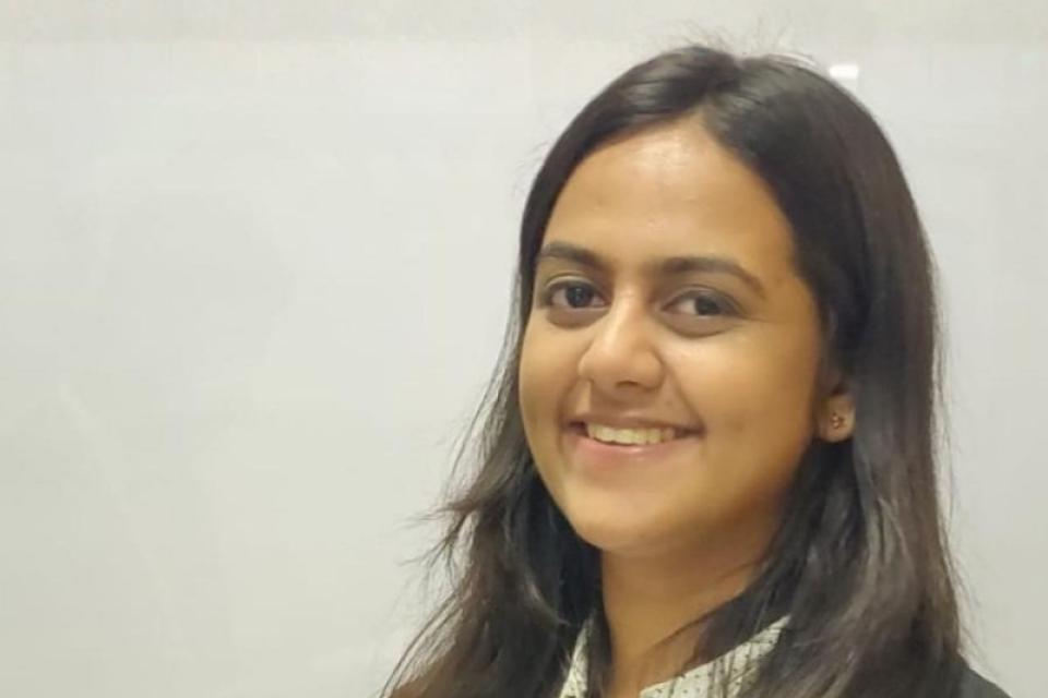 A woman with long dark hair, wearing a white blouse and black blazer, smiles while standing in front of a plain light-colored background.