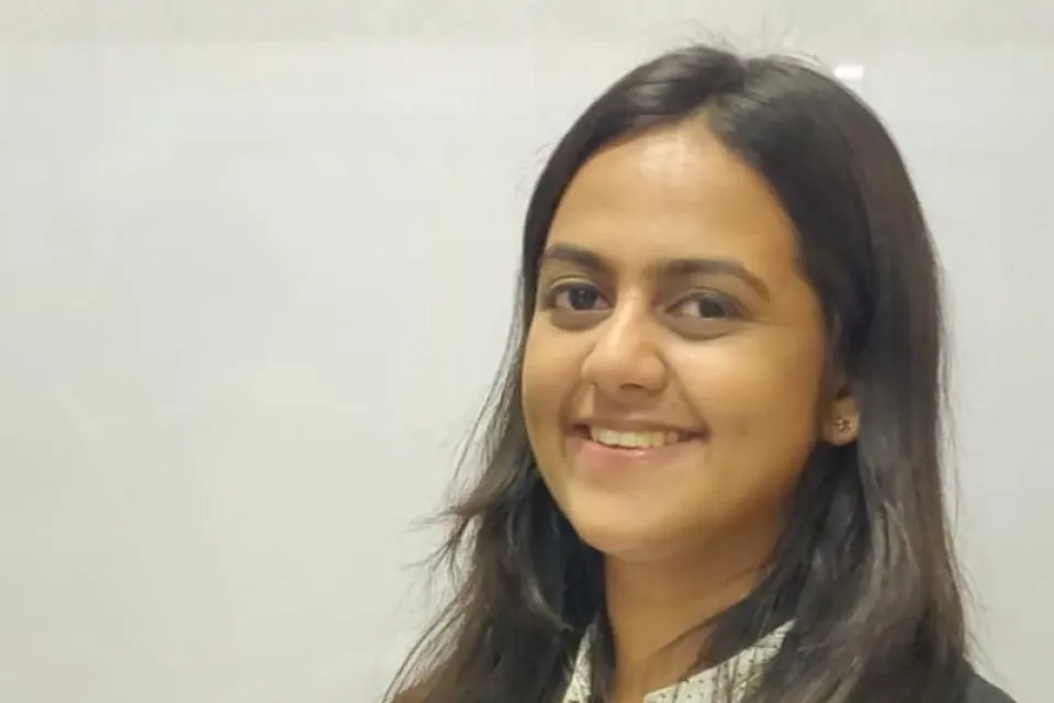 A woman with long dark hair, wearing a white blouse and black blazer, smiles while standing in front of a plain light-colored background.