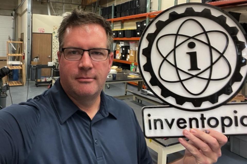 A man in a workshop holds a large sign with an atomic symbol and the word “inventopia.” Shelves and tools are visible in the background.