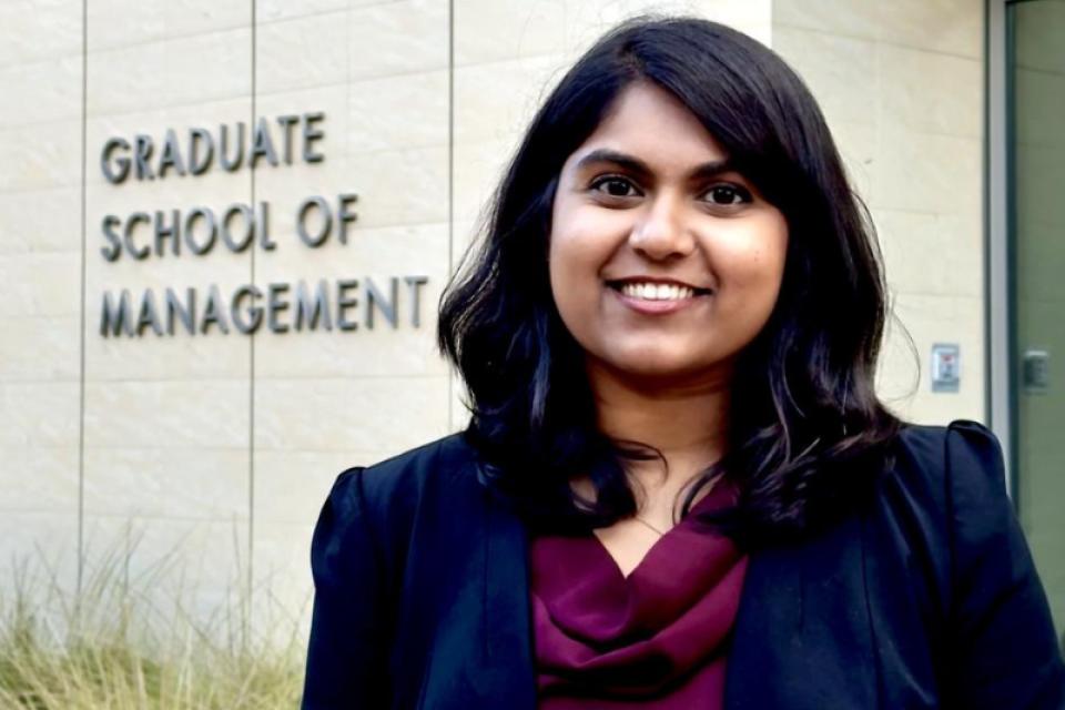 A woman in business attire stands smiling in front of a building with a sign that reads "Graduate School of Management.