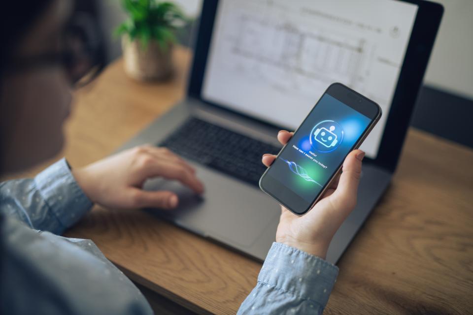 Person holding a smartphone displaying a digital assistant app icon, sitting in front of an open laptop on a desk.