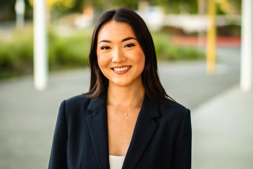 A woman in a dark blazer stands outdoors, smiling at the camera, with a blurred background of greenery and architectural structures.