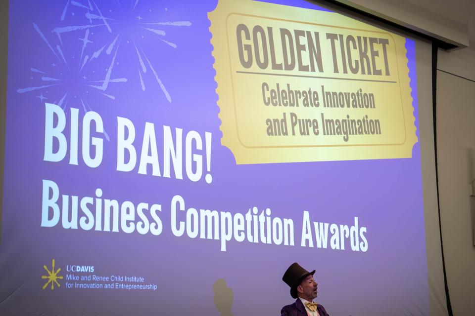 A person in a Willy Wonka costume stands in front of a projection reading "BIG BANG! Business Competition Awards" and "GOLDEN TICKET Celebrate Innovation and Pure Imagination.