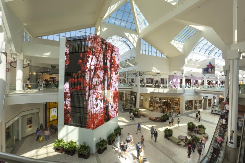 Spacious, modern shopping mall interior with a glass ceiling, digital display, multiple stores, and people walking on two levels.