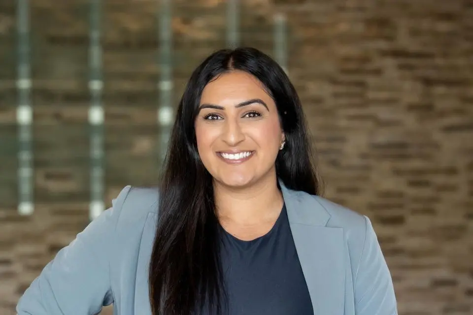 A woman with long dark hair wearing a light gray blazer and dark top stands smiling in front of a blurred brick wall background.