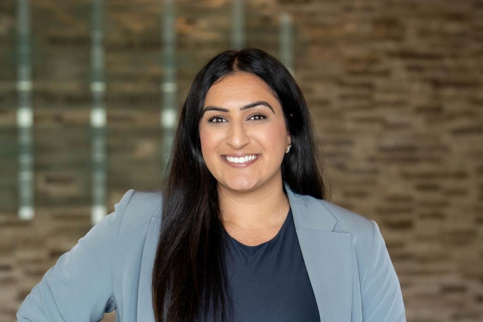 A woman with long dark hair wearing a light gray blazer and dark top stands smiling in front of a blurred brick wall background.