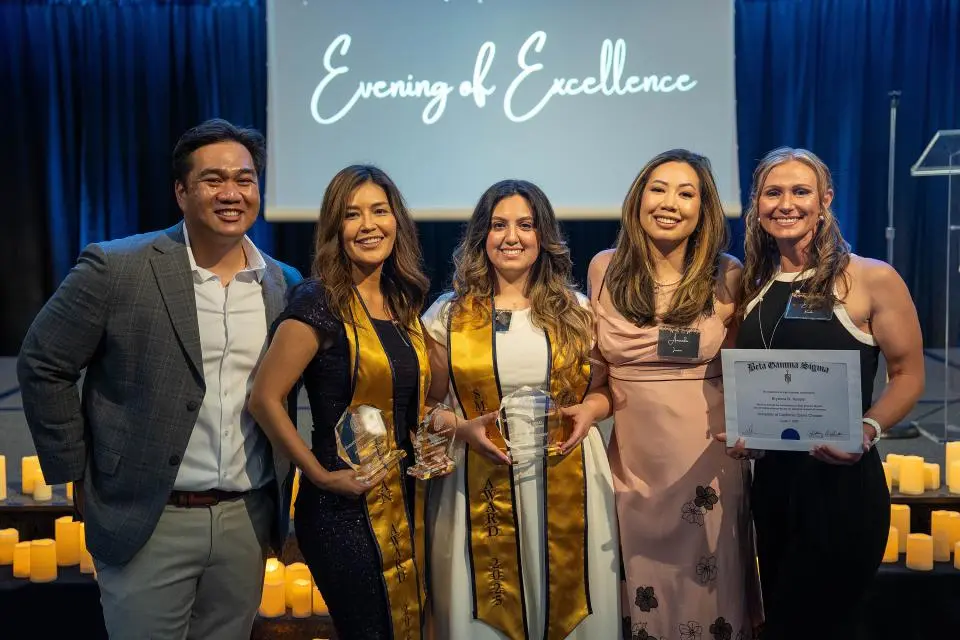 Five people pose together at an awards event; three hold glass trophies, one holds a certificate, with "Evening of Excellence" displayed on a screen behind them.