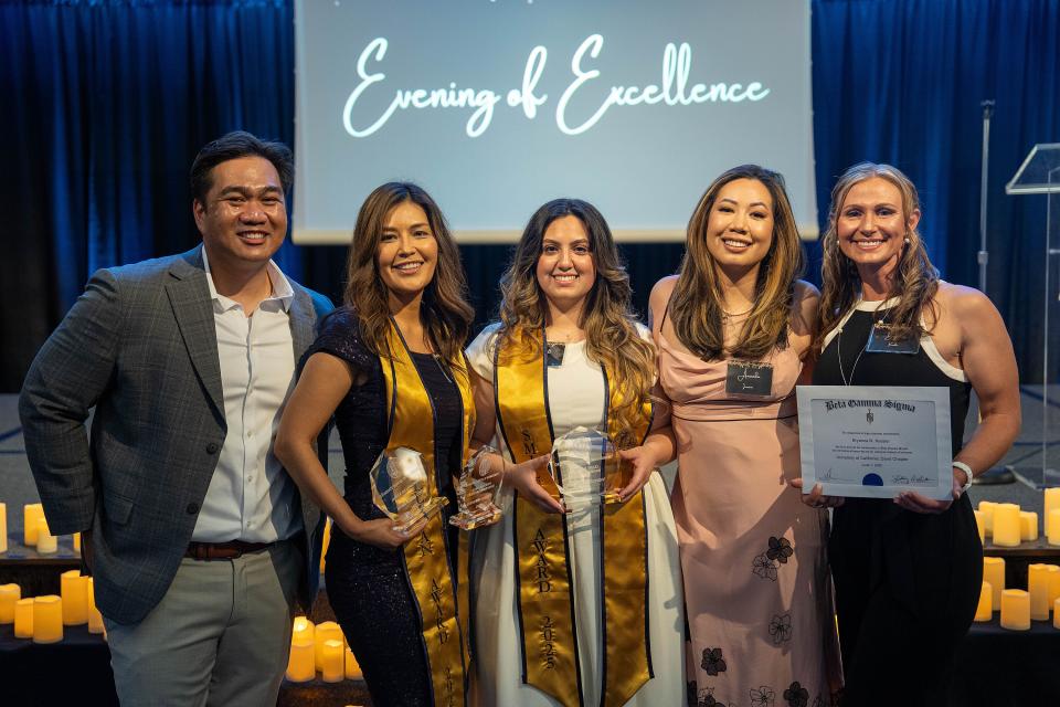 Five people pose together at an awards event; three hold glass trophies, one holds a certificate, with "Evening of Excellence" displayed on a screen behind them.