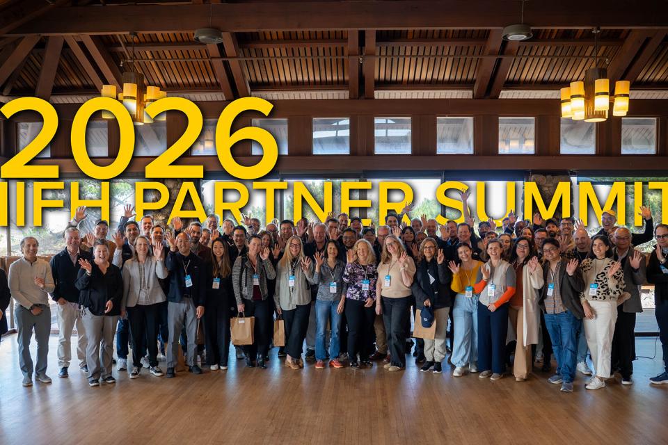 A large group of people standing together indoors, smiling and waving, with “2026 IIFH Partner Summit” in large yellow text above them.