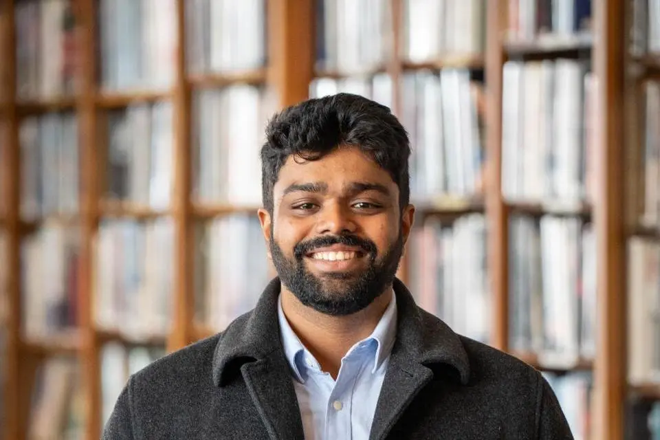 A man with dark hair and a beard, wearing a dark coat and light shirt, stands smiling in front of bookshelves filled with books.