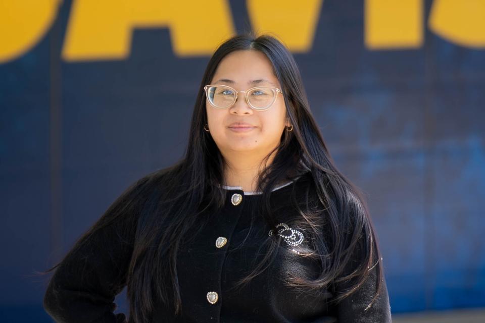Jenny Nguyen stands in front of a blue and yellow background, wearing a black sweater with decorative buttons.