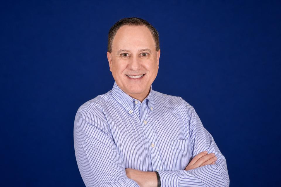 Tim Akin in a light blue striped shirt stands with arms crossed and smiles at the camera, set against a plain dark blue background.