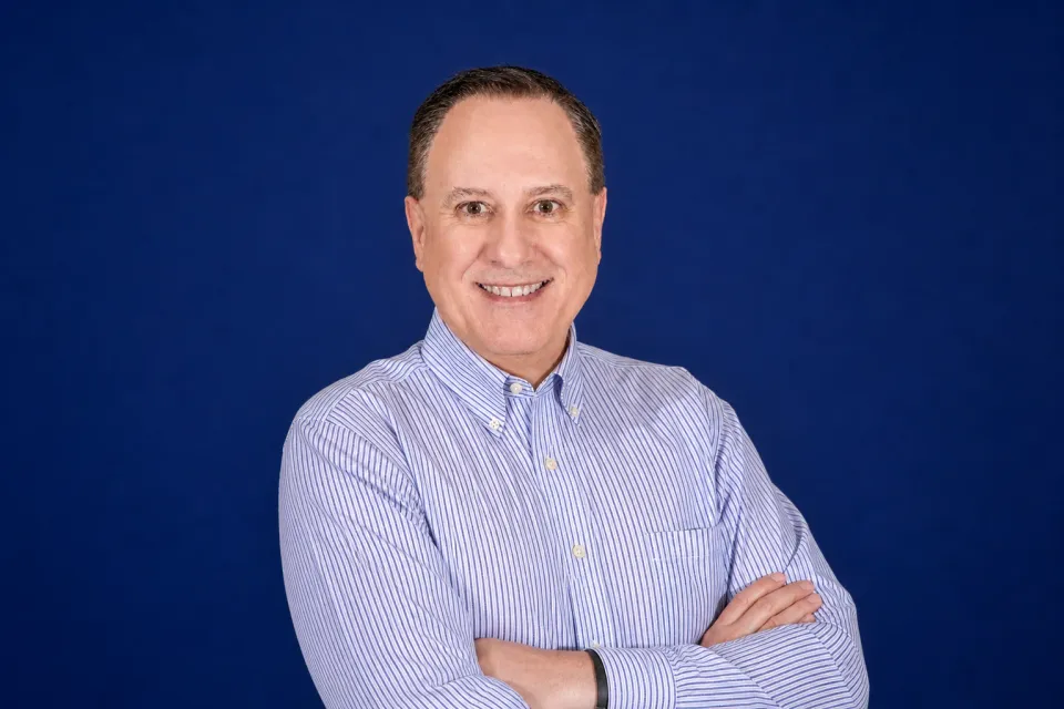 Tim Akin in a light blue striped shirt stands with arms crossed and smiles at the camera, set against a plain dark blue background.