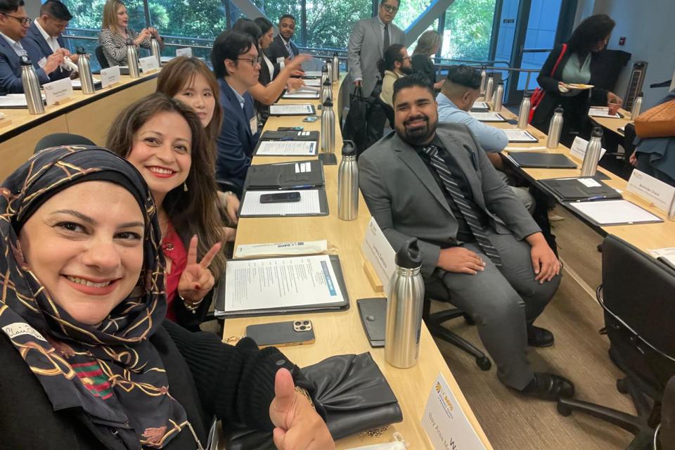 People sit in a modern classroom with large windows, facing forward. Some smile and pose for a photo, while others talk or look at materials on the desks.