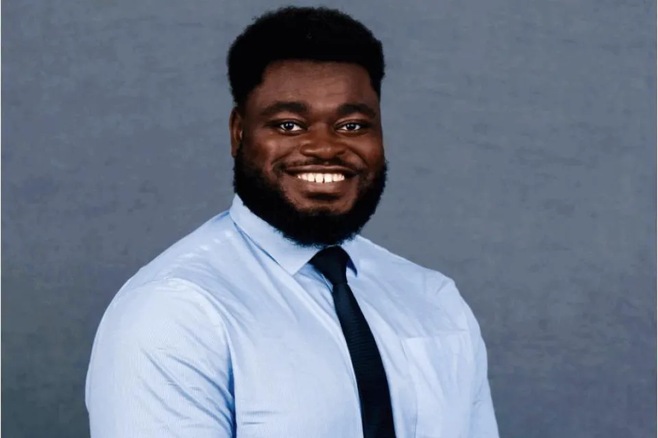 A man with a beard and afro hairstyle, wearing a light blue dress shirt and dark tie, smiles in front of a plain gray background.