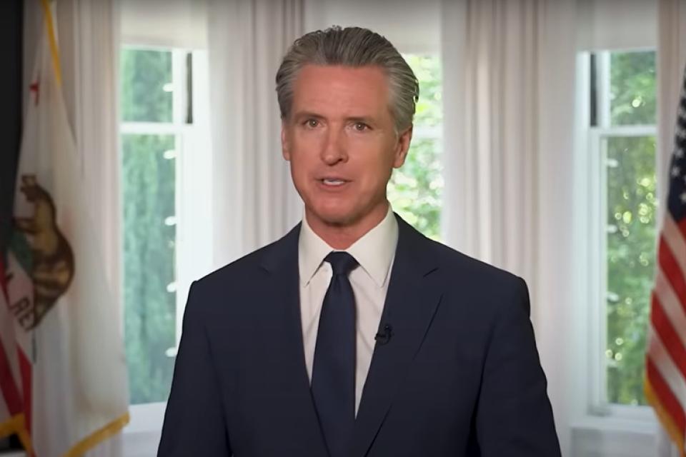 Gavin Newsom in a navy suit and tie stands indoors between two windows, with American and California state flags visible in the background.