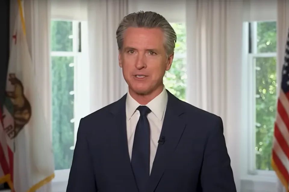 Gavin Newsom in a navy suit and tie stands indoors between two windows, with American and California state flags visible in the background.