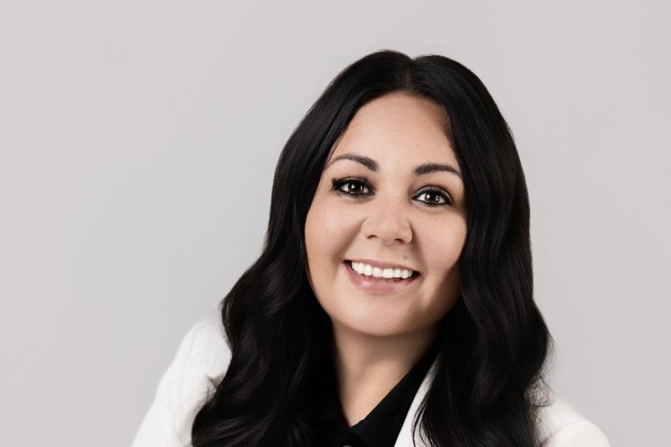A woman with long dark hair, wearing a white blazer over a black top, is seated and smiling at the camera against a plain light background.
