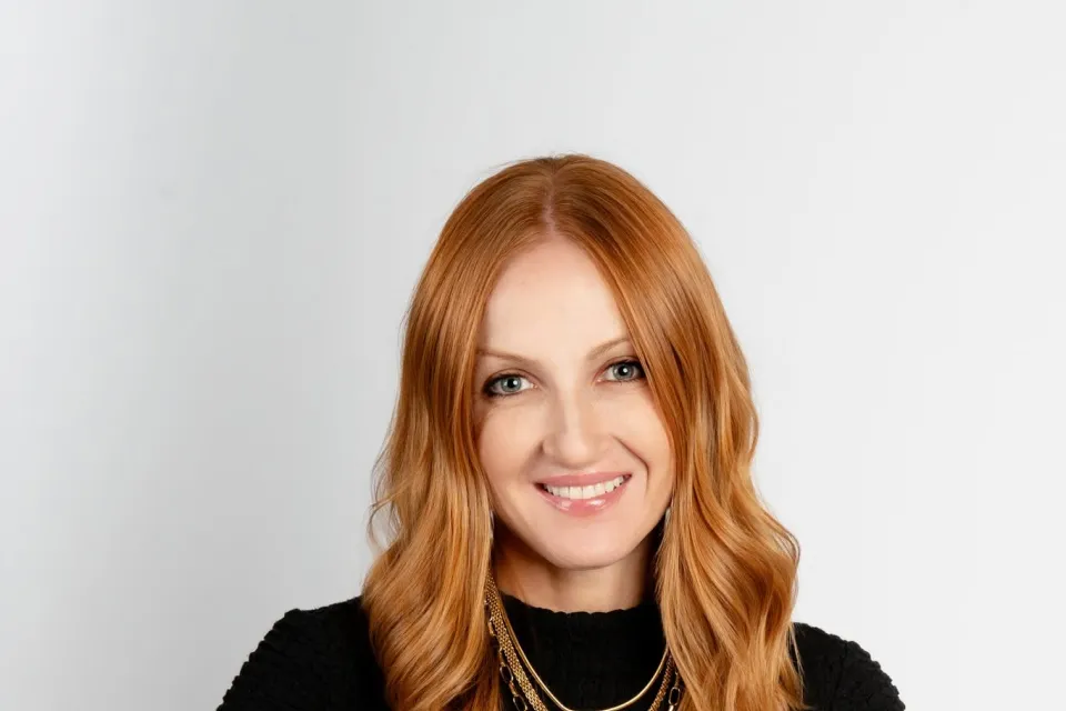 Woman with long red hair wearing a black textured top and layered gold necklaces, seated and smiling against a plain light background.