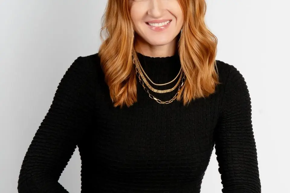 Woman with long red hair wearing a black textured top and layered gold necklaces, seated and smiling against a plain light background.